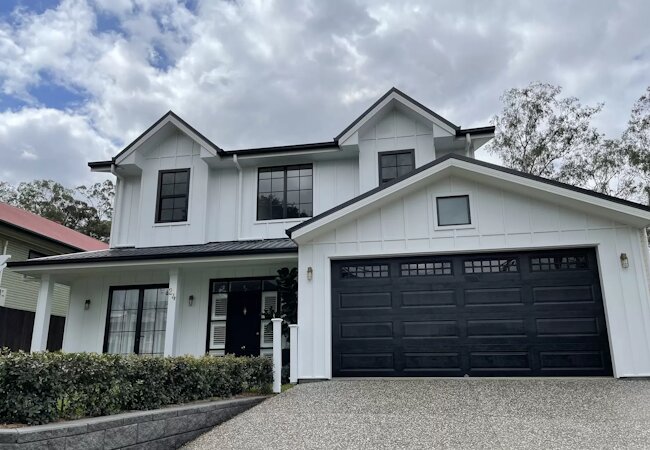 Modern two-story white house with black garage door, black window frames, and a gravel driveway under a cloudy sky.
