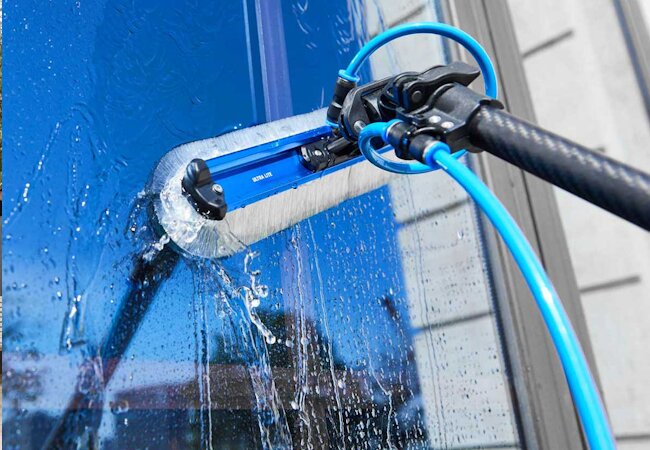 Close-up of a water-fed pole brush cleaning a blue window with water flowing down the glass.