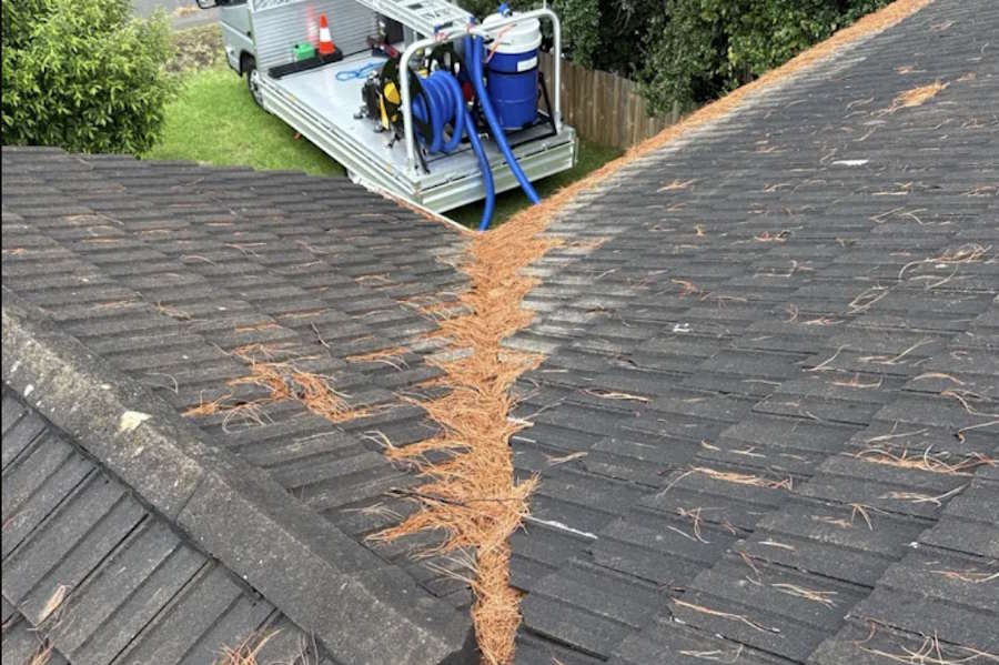 Roof valley with scattered pine needles and a truck with cleaning equipment parked on grass nearby.