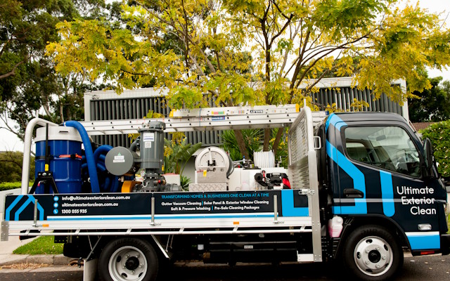 Utility truck equipped with cleaning equipment and ladders, branded with Ultimate Exterior Clean, parked on a street with leafy trees in the background.