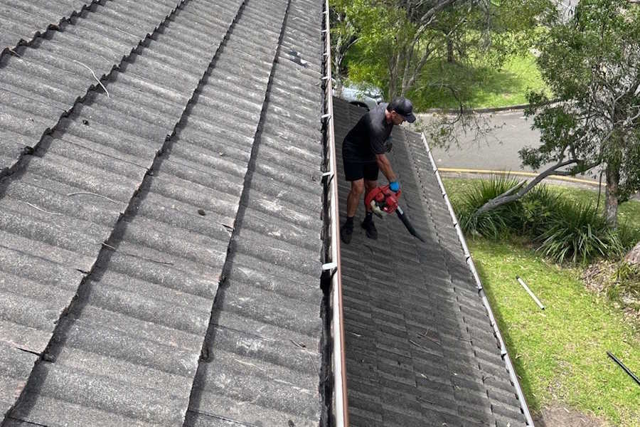 Man in black clothes using a leaf blower to clean a tiled roof near a gutter surrounded by green trees and grass.